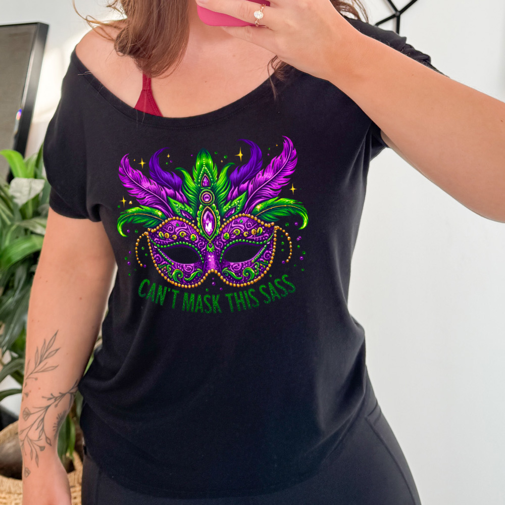 black slouchy tee with the graphic of a fancy, feathered and bejeweled, and colorful mardi gras mask, and words underneath saying "can't mask this sass"