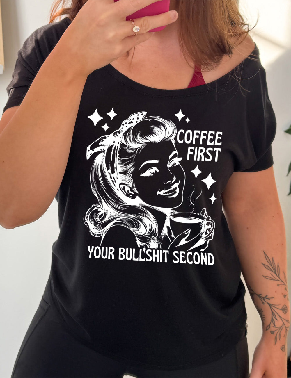 black slouchy tee with the graphic of a woman happily holding a mug of steaming coffee, with the words "coffee first" (next to her face) and "your bullshit second" underneath, and a few stars around her