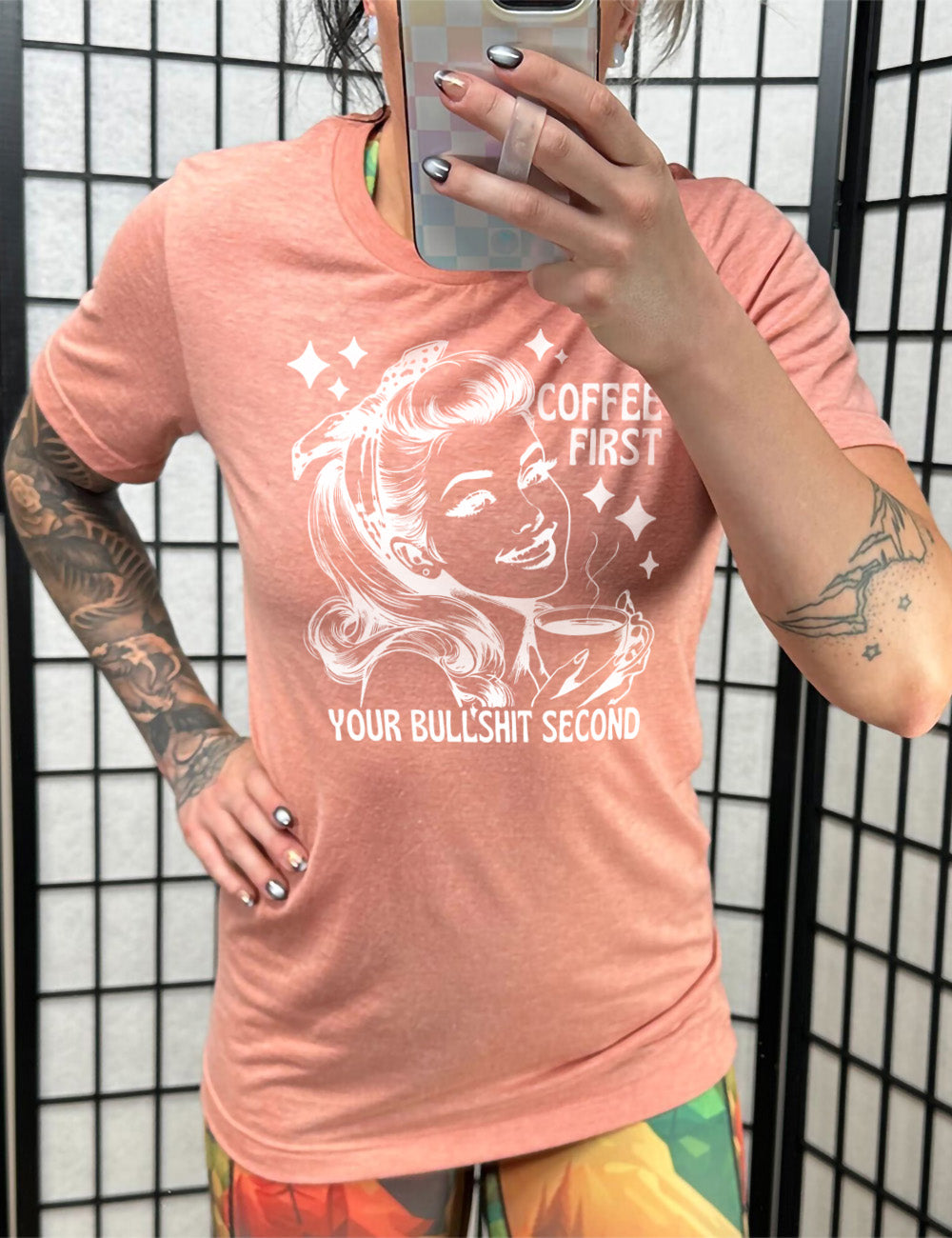 sunset unisex tee with the graphic of a woman happily holding a mug of steaming coffee, with the words "coffee first" (next to her face) and "your bullshit second" underneath, and a few stars around her