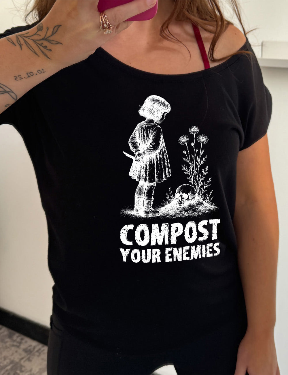 black slouchy tee with the graphic of a little bobbed-hair girl in a dress, holding a knife behind her back, looking down at a skull that rests on a little hill with tall flowers growing there, and the words underneath that say "compost your enemies"