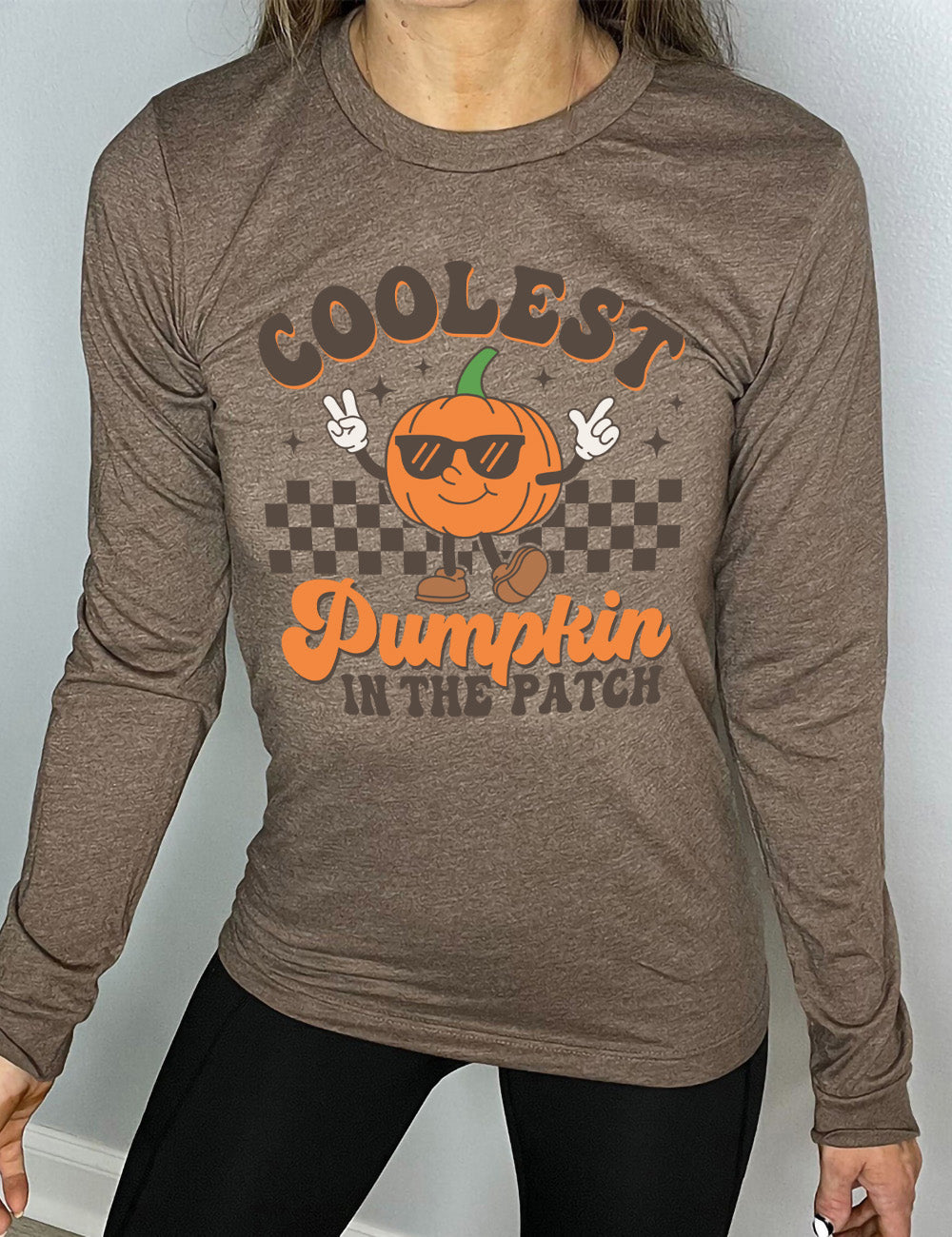 heather brown long sleeve with the graphic of a pumpkin, wearing sunglasses, both arms raised, foot kicking out, in front of a checkered banner, surrounded by a few little stars, with the words around it saying "coolest pumpkin in the patch"