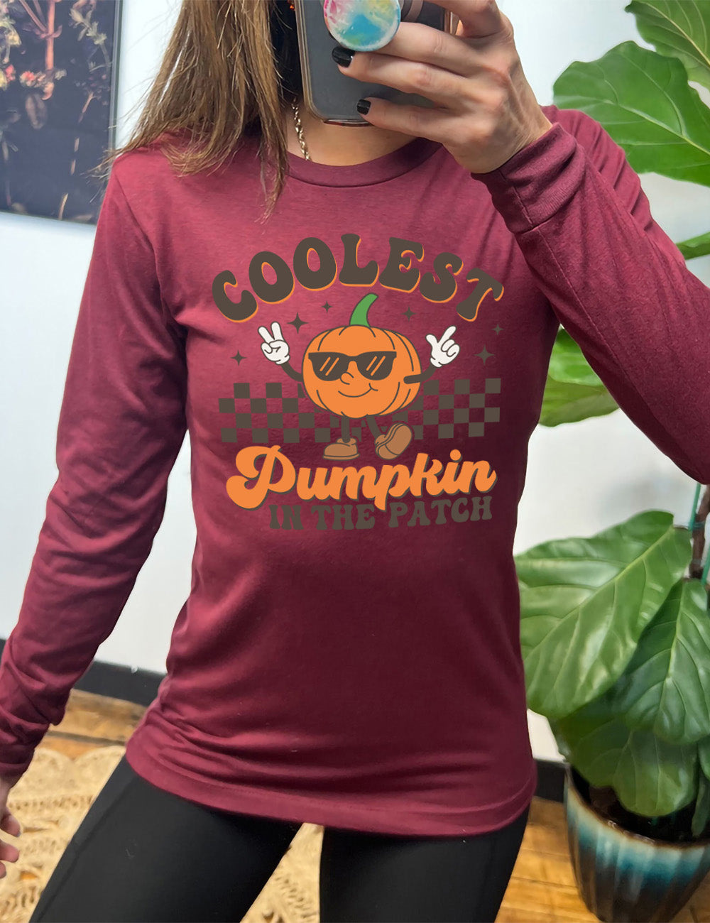 heather cardinal long sleeve with the graphic of a pumpkin, wearing sunglasses, both arms raised, foot kicking out, in front of a checkered banner, surrounded by a few little stars, with the words around it saying "coolest pumpkin in the patch"