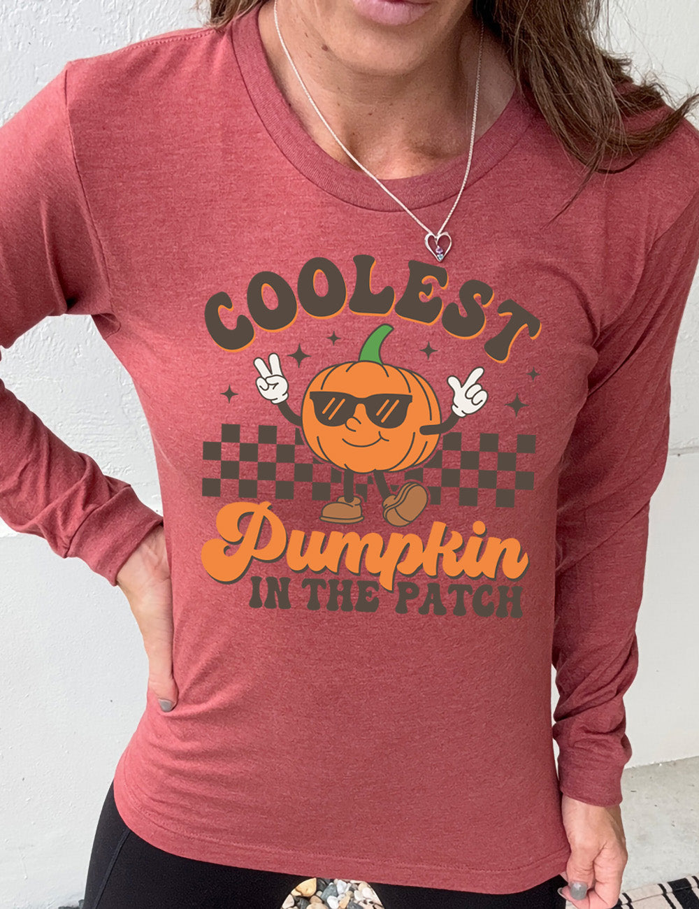 heather clay long sleeve with the graphic of a pumpkin, wearing sunglasses, both arms raised, foot kicking out, in front of a checkered banner, surrounded by a few little stars, with the words around it saying "coolest pumpkin in the patch"