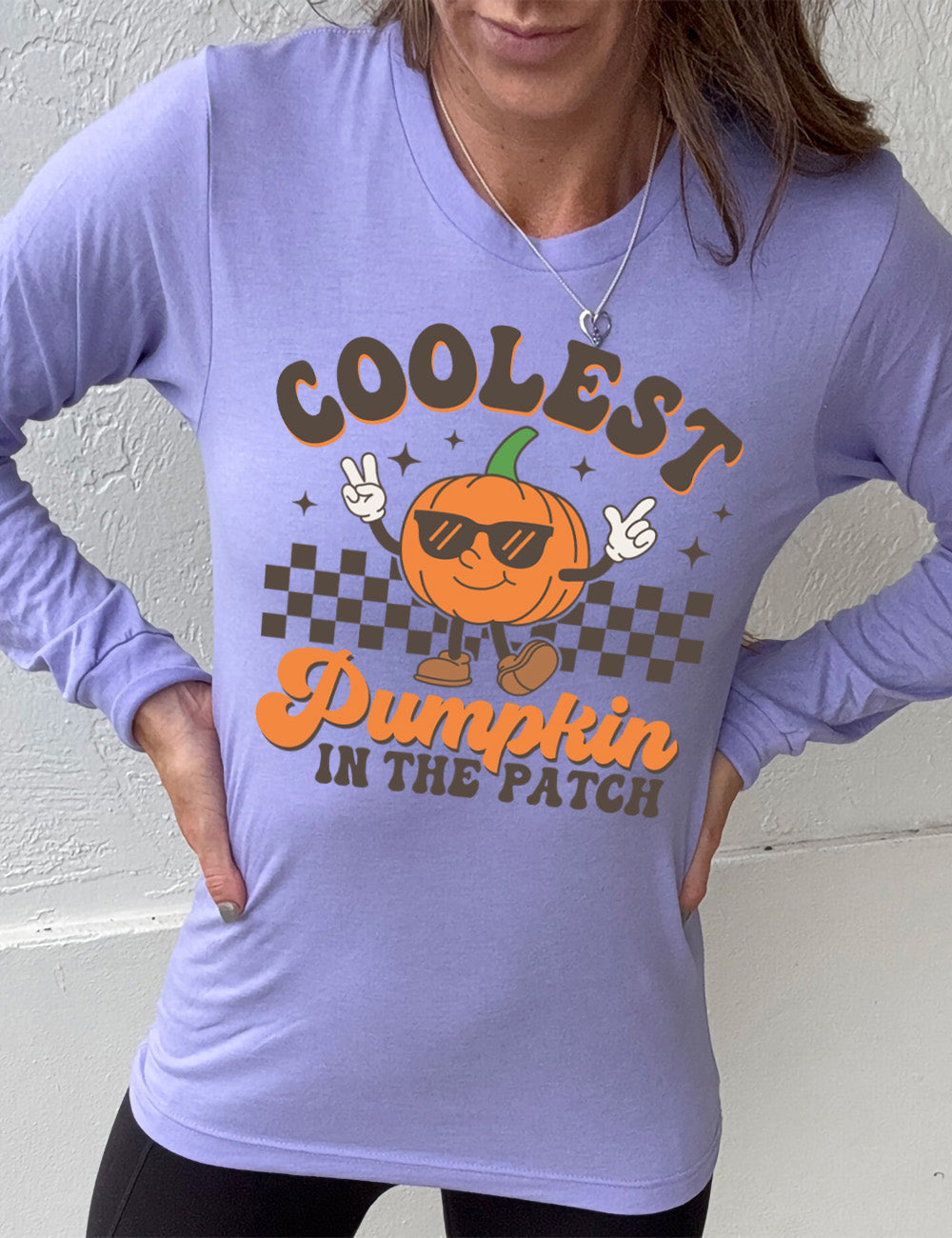 heather dark lavender long sleeve with the graphic of a pumpkin, wearing sunglasses, both arms raised, foot kicking out, in front of a checkered banner, surrounded by a few little stars, with the words around it saying "coolest pumpkin in the patch"