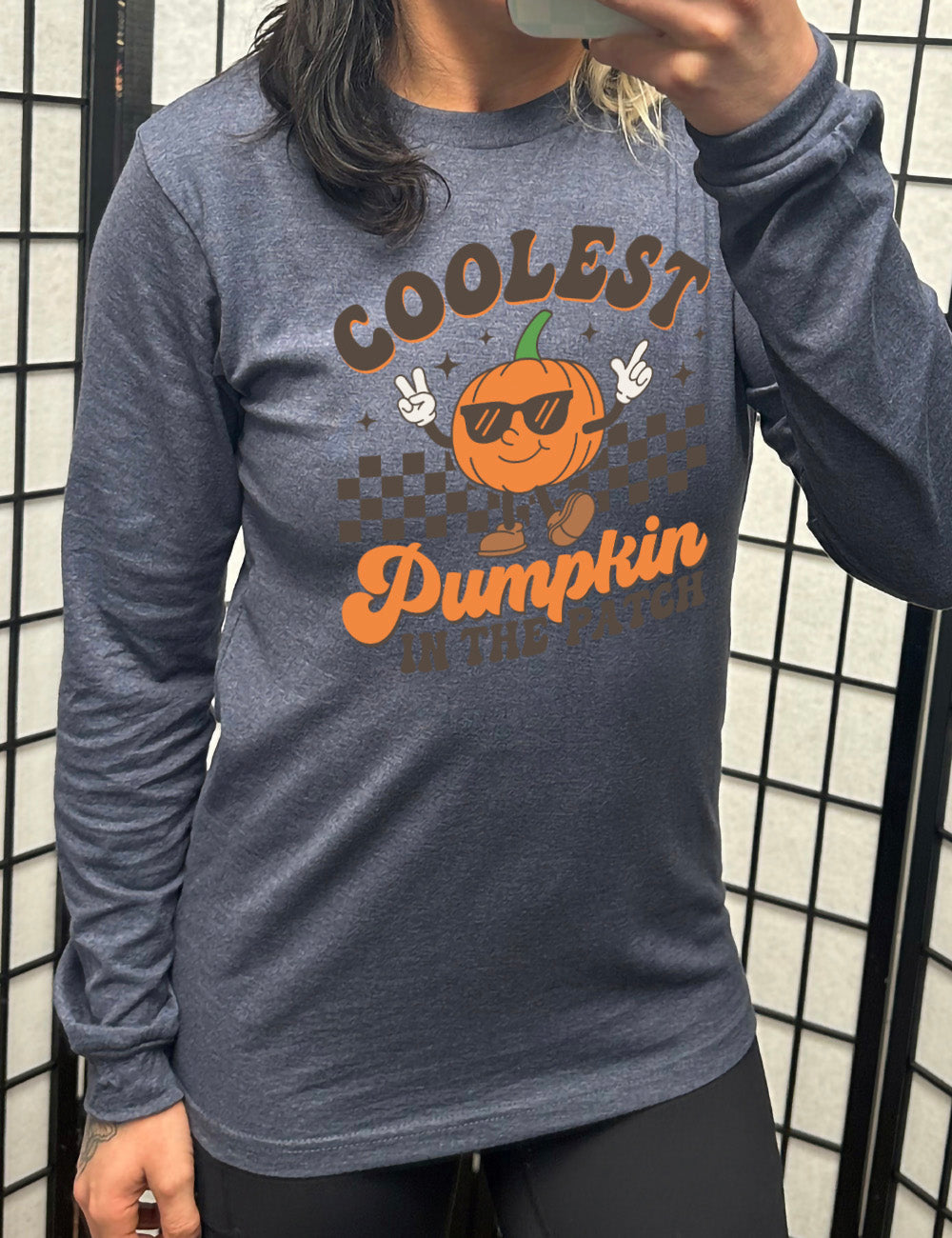 heather navy long sleeve with the graphic of a pumpkin, wearing sunglasses, both arms raised, foot kicking out, in front of a checkered banner, surrounded by a few little stars, with the words around it saying "coolest pumpkin in the patch"
