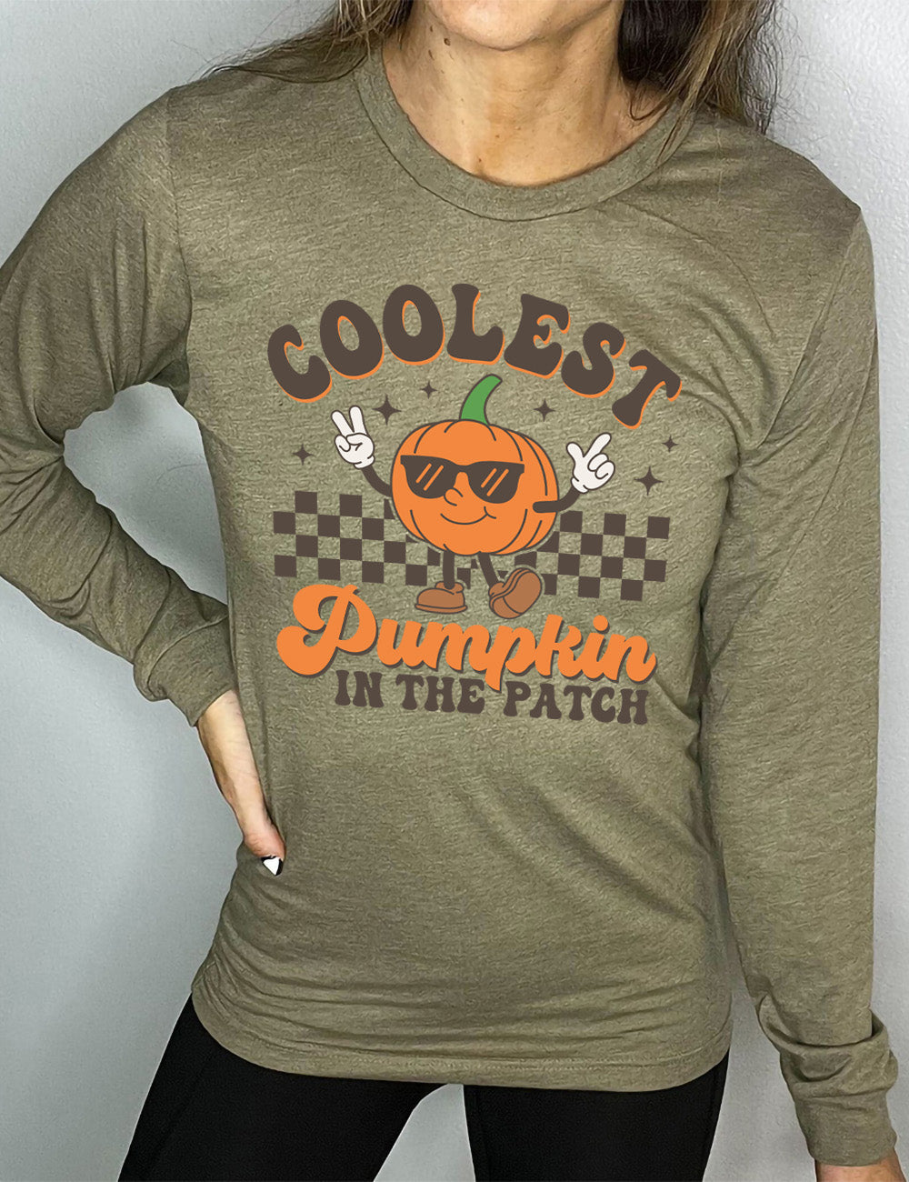 heather olive long sleeve with the graphic of a pumpkin, wearing sunglasses, both arms raised, foot kicking out, in front of a checkered banner, surrounded by a few little stars, with the words around it saying "coolest pumpkin in the patch"