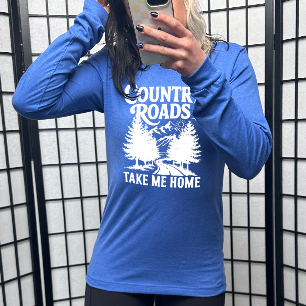 heather true royal long sleeve with the words "country roads take me home" on it, and the graphic of a road going through tall trees, with mountains beyond, and the sun