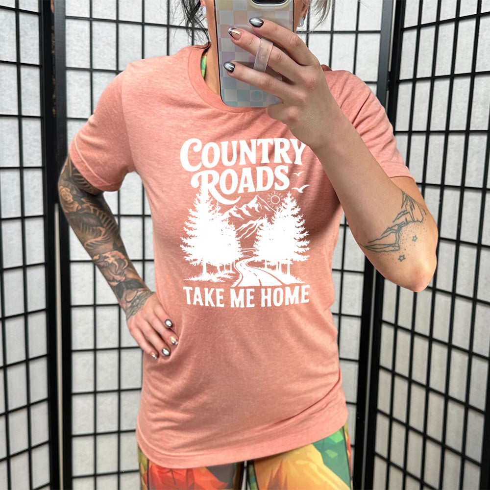 sunset unisex tee with the words "country roads take me home" on it, and the graphic of a road going through tall trees, with mountains beyond, and the sun