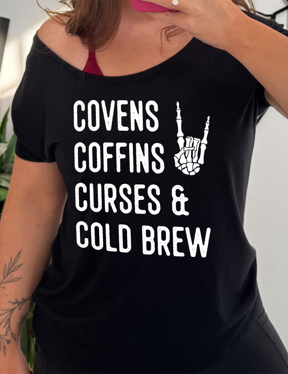 black slouchy tee with the words listed "covens coffins curses & cold brew" and the graphic of a skeletal rocker hand