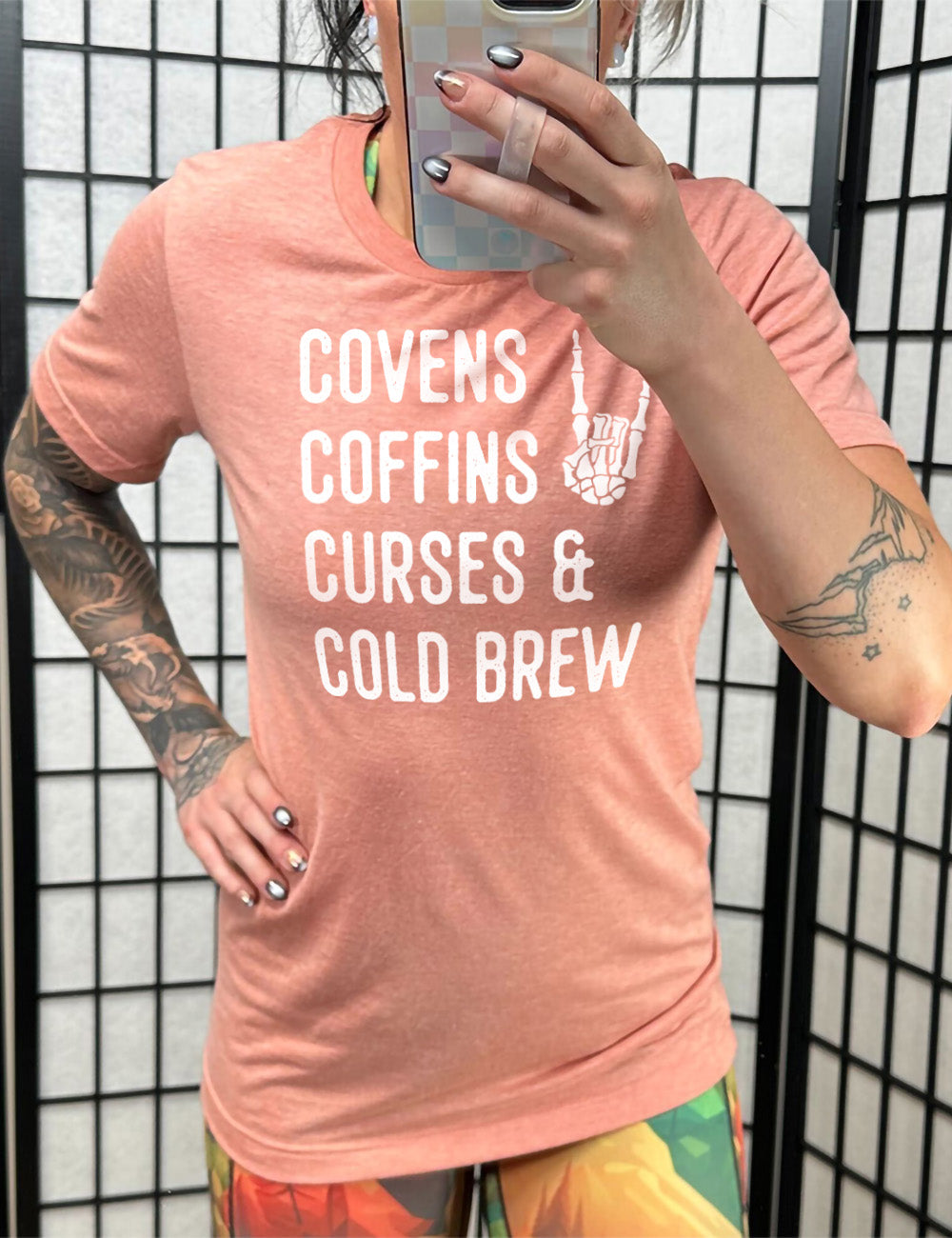 sunset unisex tee with the words listed "covens coffins curses & cold brew" and the graphic of a skeletal rocker hand