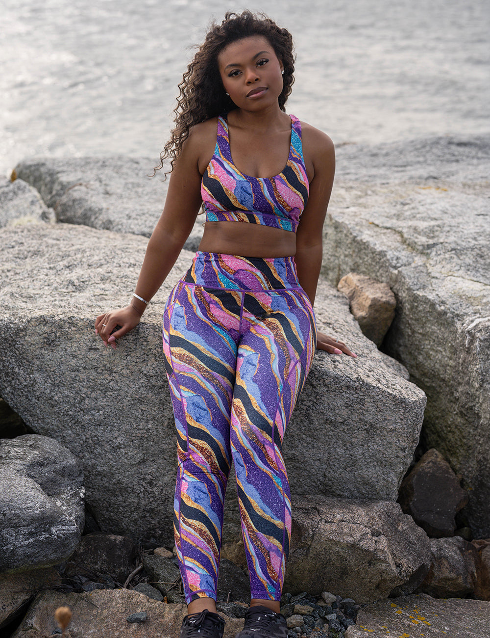 model wearing the purple, gold, black, blue, and pink abstract stripes full length leggings and sports bra at the ocean
