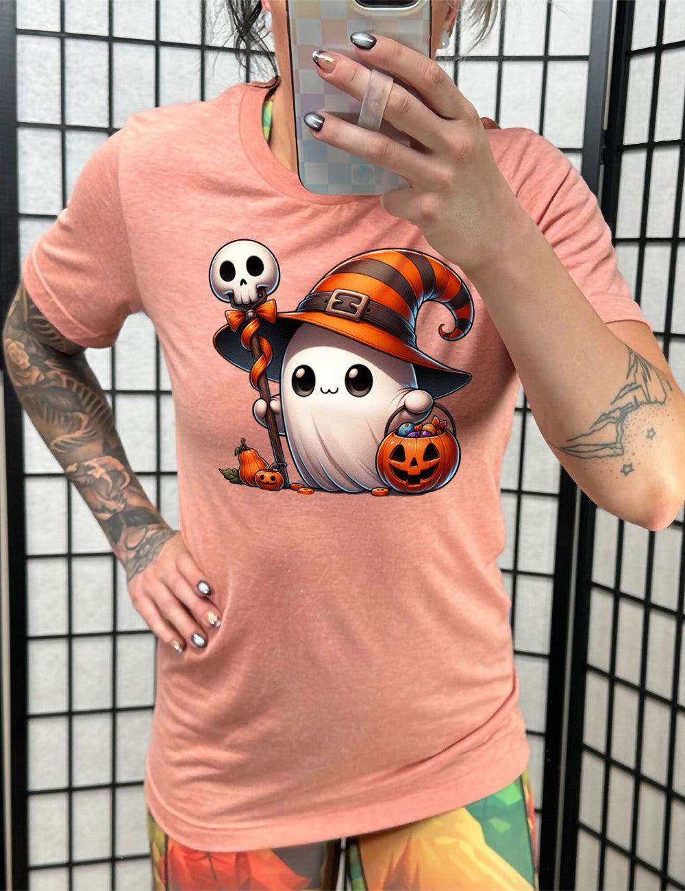 sunset unisex tee with the graphic of a cute ghost wearing a striped witch hat, holding a candy-filly jack-o-lantern basket as well as a staff with a skull on top and a bow with ribbon, all next to a tiny gourd and a jack-o-lantern
