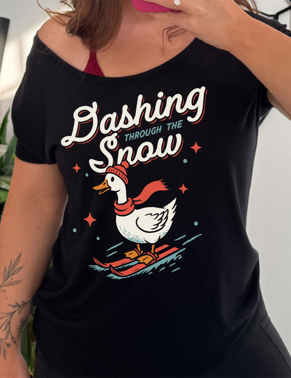black slouchy tee with the graphic of a goose, wearing a hat and scarf, on skis, in  downwards motion,  with some stars, and the words above it saying "Dashing through the Snow" 