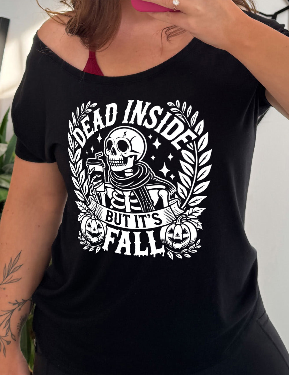 black slouchy tee with the words "dead inside but it's fall" on it, with plants framing the sides, two jack-o-lantern graphics, and the large graphic in the middle of a skeleton, wearing a scarf and holding a takeaway drink with a straw, and a few stars