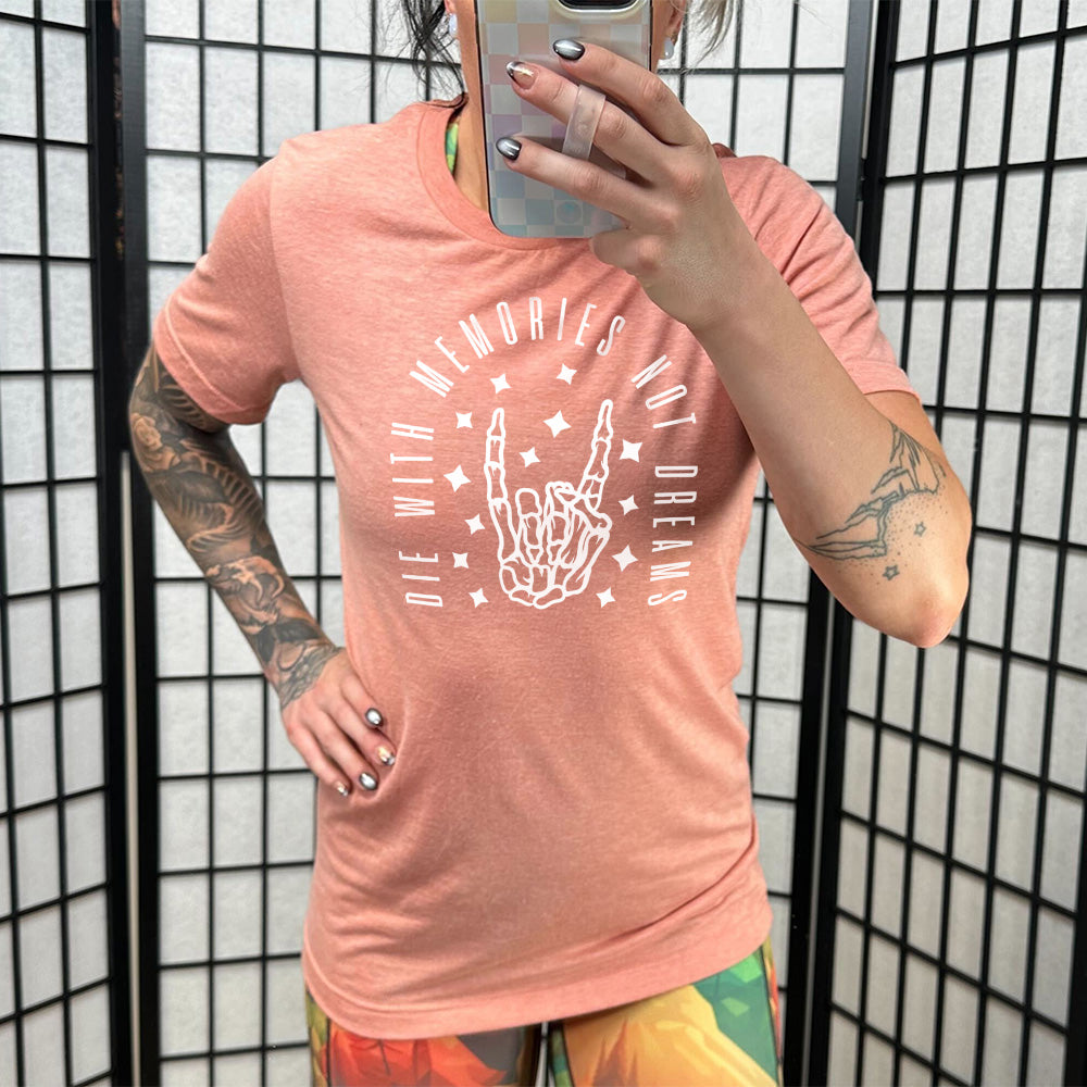 sunset unisex tee with the graphic of a rocker skeleton hand, surrounded by little stars as well as an archway of text saying "die with memories not dreams" 