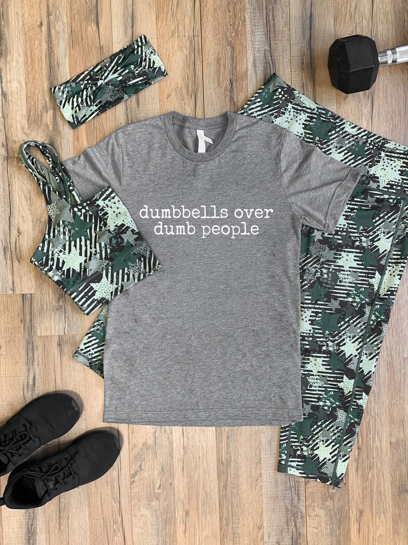 A flat-lay of a women's activewear set featuring a heather gray t-shirt with the text 'dumbbells over dumb people' in white, paired with green and black camouflage leggings, a matching headband, a sports bra, and black sneakers, arranged on a wooden floor next to a dumbbell.