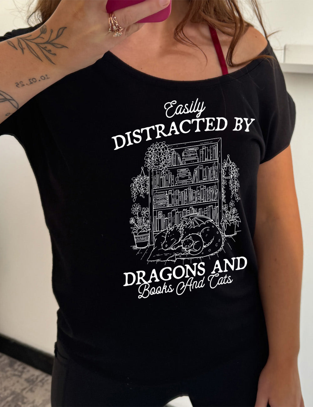 black slouchy tee with words on it saying "Easily distracted by dragons and books and cats" around the graphic of a detailed large, full bookcase, with plants hanging and placed beside it, and a dragon curled up in front of it