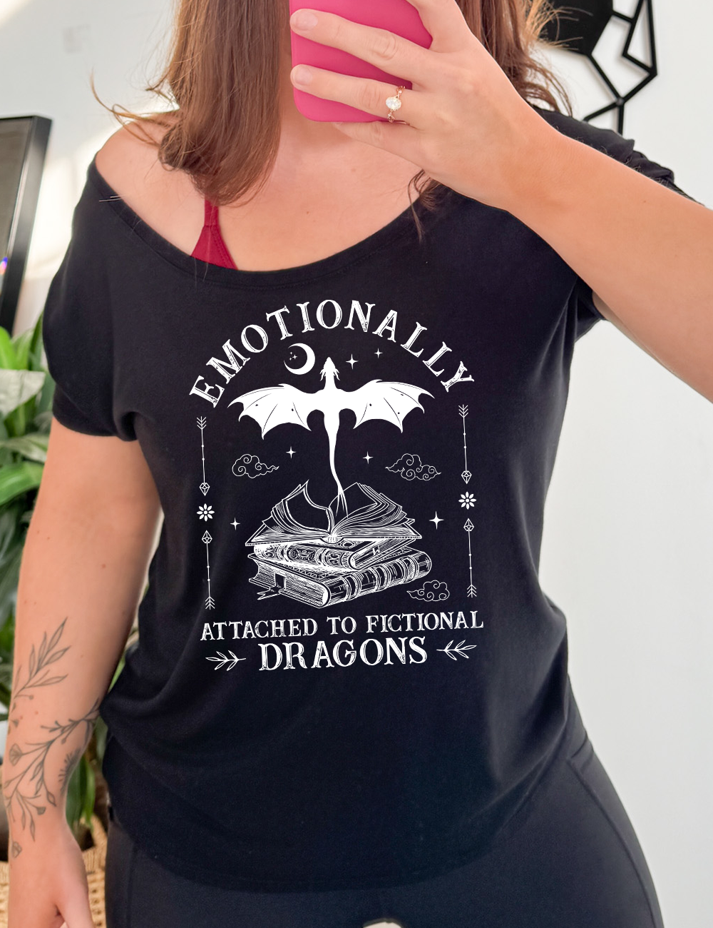 black slouchy tee with the words "emotionally attached to fictional dragons" on it, with graphics of a stack of books, with a dragon flying out of the open book on top, few a few little clouds, a moon, some stars, and arrows and flowers