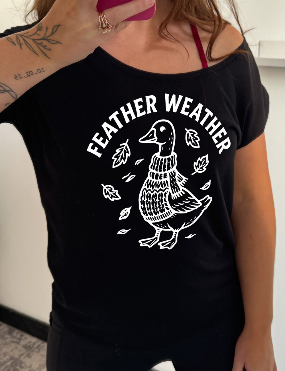 black slouchy tee with the graphic of a goose, wearing a scarf and sweater, surrounded by falling leaves, and the words arched over it saying "feather weather"