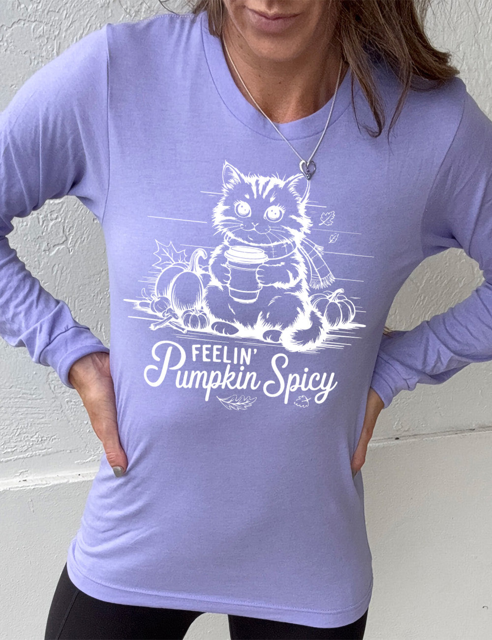 heather dark lavender long sleeve with the graphic of a cat, wearing a scarf, next to a few pumpkins of various sizes and a few falling leaves, holding a takeaway coffee, and the words underneath saying "feelin' pumpkin spicy"