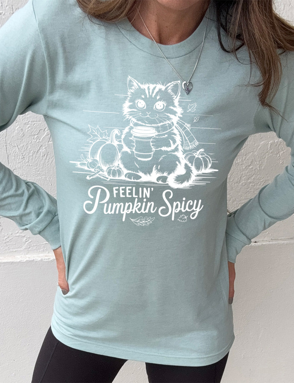 heather dusty blue long sleeve with the graphic of a cat, wearing a scarf, next to a few pumpkins of various sizes and a few falling leaves, holding a takeaway coffee, and the words underneath saying "feelin' pumpkin spicy"