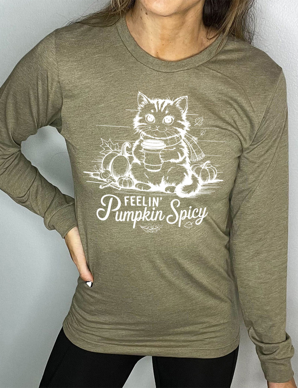 heather olive long sleeve with the graphic of a cat, wearing a scarf, next to a few pumpkins of various sizes and a few falling leaves, holding a takeaway coffee, and the words underneath saying "feelin' pumpkin spicy"