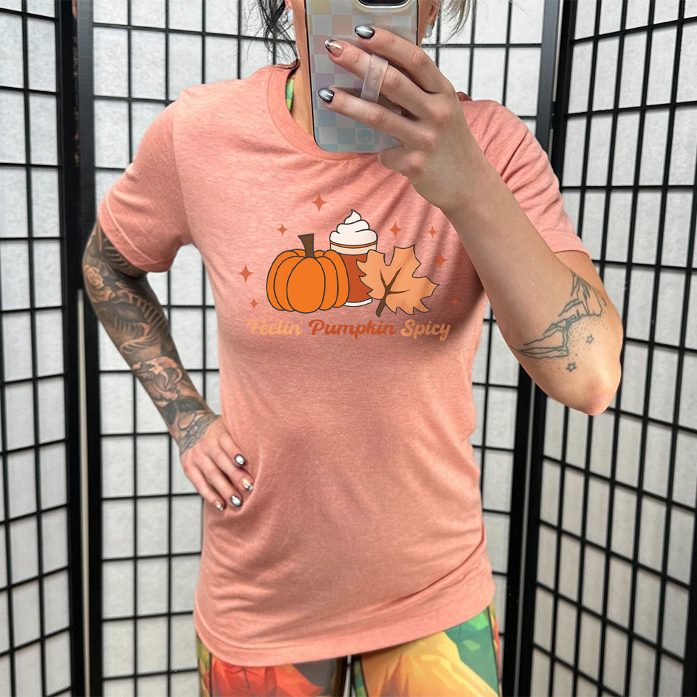 Model wearing sunset colored shirt with pumpkin, coffee, and fall leaf graphic on it that says "Feelin' Pumpkin Spicy"
