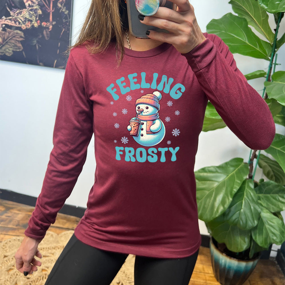 heather cardinal long sleeve shirt with the words "feeling frosty" on it, with the graphic of snowperson, wearing a hat and scarf, drinking an iced drink, surrounded by snowflakes