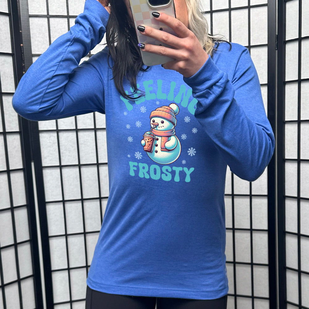 heather true royal long sleeve shirt with the words "feeling frosty" on it, with the graphic of snowperson, wearing a hat and scarf, drinking an iced drink, surrounded by snowflakes