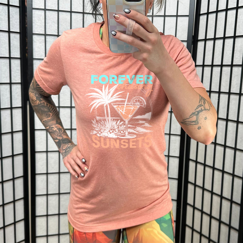 Model wearing sunset colored shirt with a drink on the beach graphic on it that says "Forever Chasing Sunsets" 