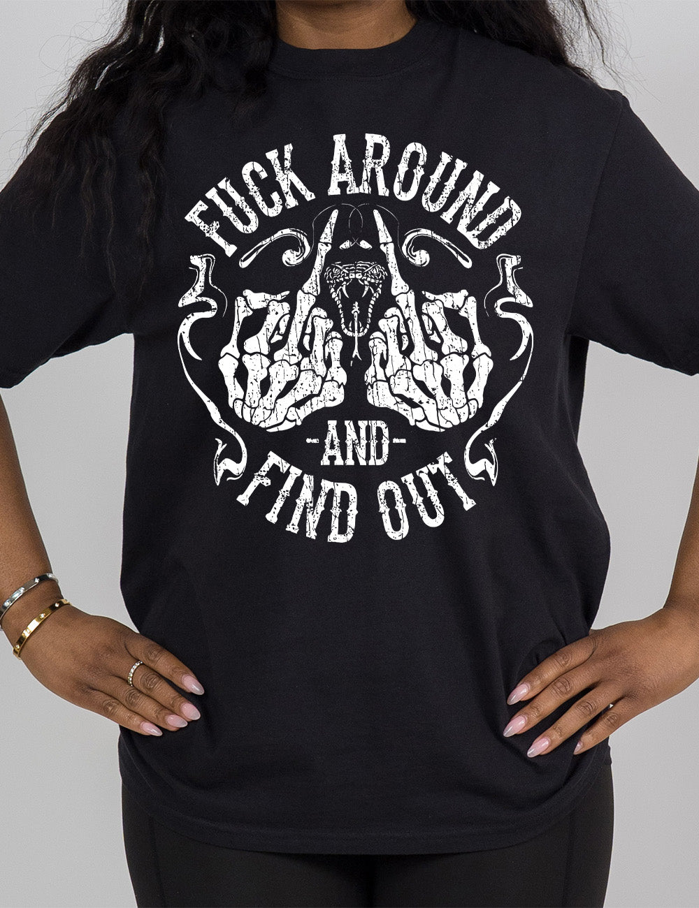 black modern tee with the graphic of two skeletal hands both giving the middle finger, around a snake's head with its tongue out, with the words circling around this all saying "fuck around and find out" with smokey lines completing the sides of the circle
