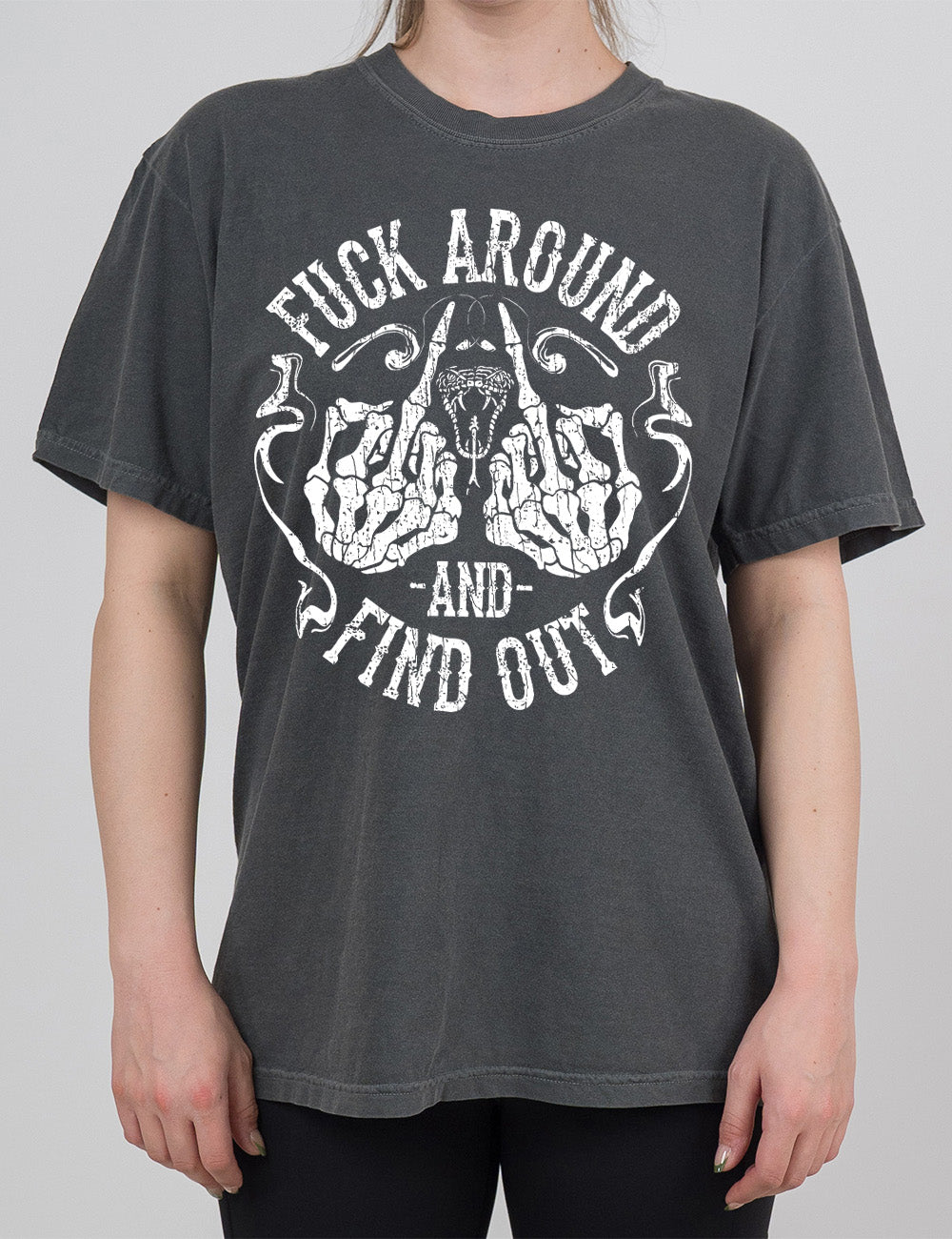 pepper modern tee with the graphic of two skeletal hands both giving the middle finger, around a snake's head with its tongue out, with the words circling around this all saying "fuck around and find out" with smokey lines completing the sides of the circle