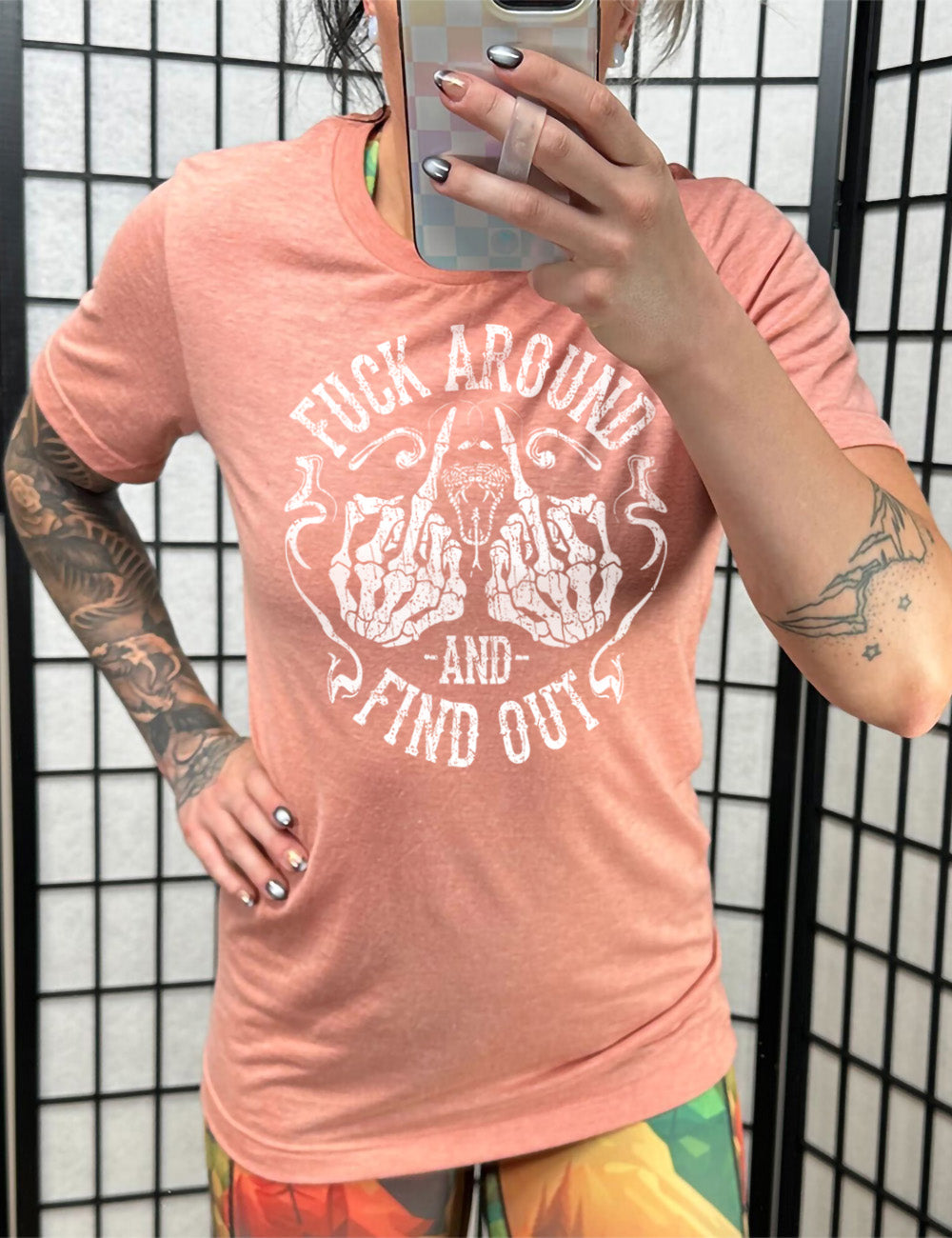 sunset unisex tee with the graphic of two skeletal hands both giving the middle finger, around a snake's head with its tongue out, with the words circling around this all saying "fuck around and find out" with smokey lines completing the sides of the circle