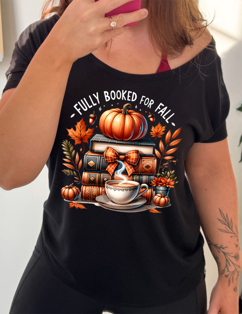 black slouchy tee with the colorful graphic of a stack of books, topped with a pumpkin, surrounded by plants and acorns and two little pumpkins, and a steaming teacup in front, with the words above saying "fully booked for fall"