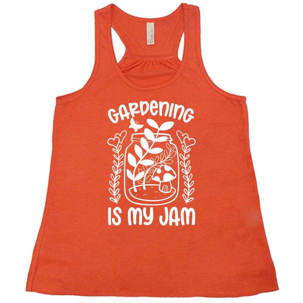 coral flowy racerback tank with the words "gardening is my jam" on it, and the graphic of a jar with plants and a mushroom in it, a little butterfly flying above it, and a plant (with hearts for flowers on them) on both sides of it