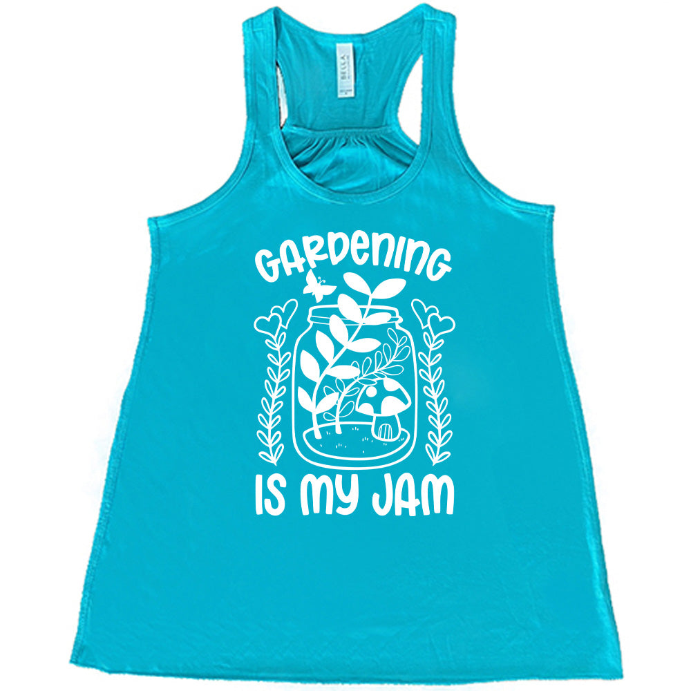 teal flowy racerback tank with the words "gardening is my jam" on it, and the graphic of a jar with plants and a mushroom in it, a little butterfly flying above it, and a plant (with hearts for flowers on them) on both sides of it