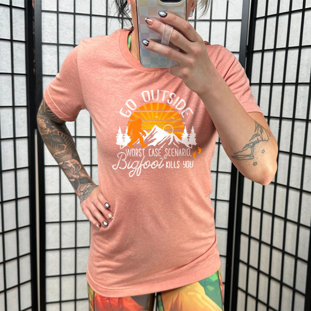 Model wearing sunset colored shirt with sun and bigfoot in mountains graphic on it that says "Go Outside, Worst Case Scenario Bigfoot Kills You"