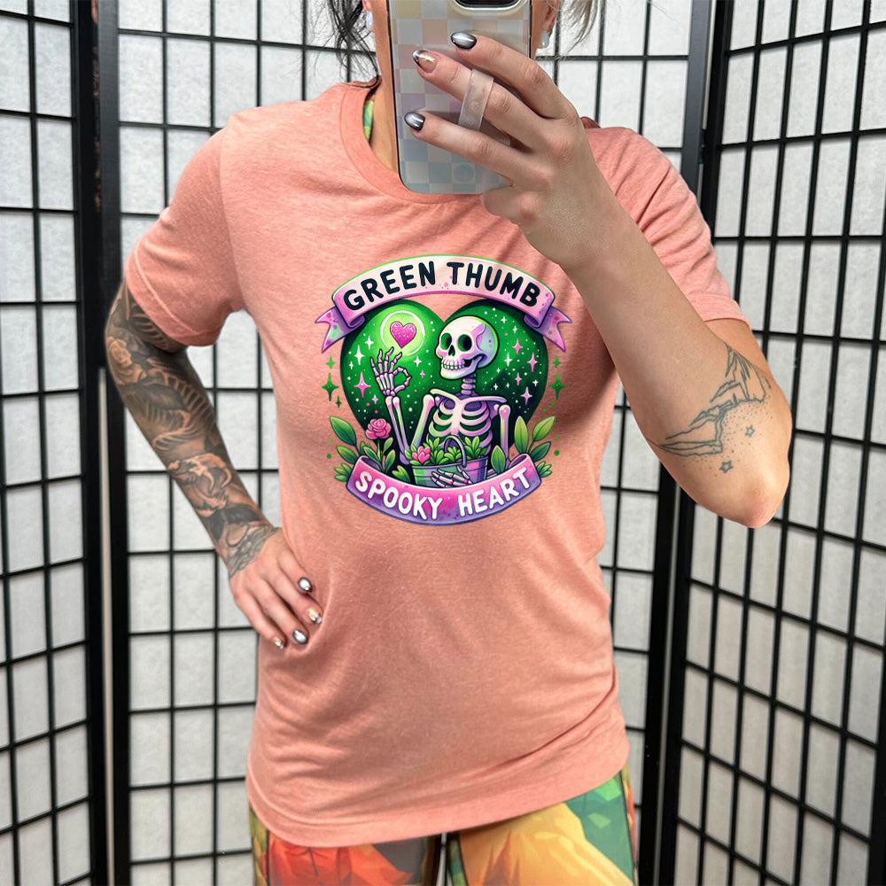 Model wearing sunset colored shirt with a skeleton and plants graphic on it that says "Green Thumb Spooky Heart"
