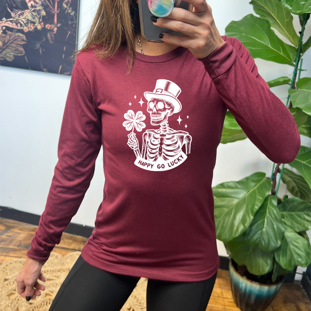 heather cardinal long sleeve shirt with the graphic of a skeleton wearing a leprechaun hat and holding a four-leaf clover, with a few little stars around, and the text "happy go lucky" underneath