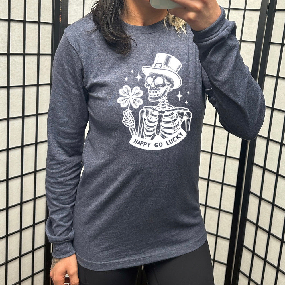 heather navy long sleeve shirt with the graphic of a skeleton wearing a leprechaun hat and holding a four-leaf clover, with a few little stars around, and the text "happy go lucky" underneath