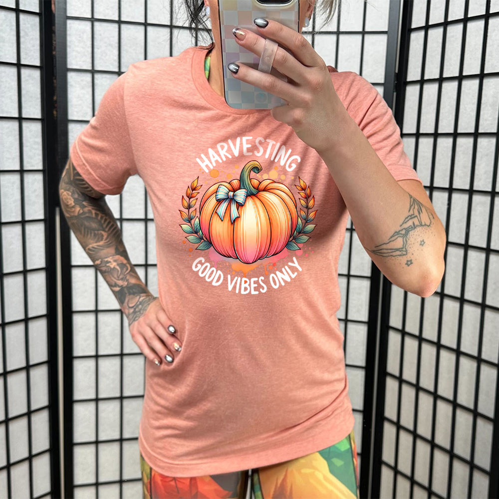 Model wearing sunset colored shirt with pumpkin graphic on it that says "Harvesting Good Vibes Only"