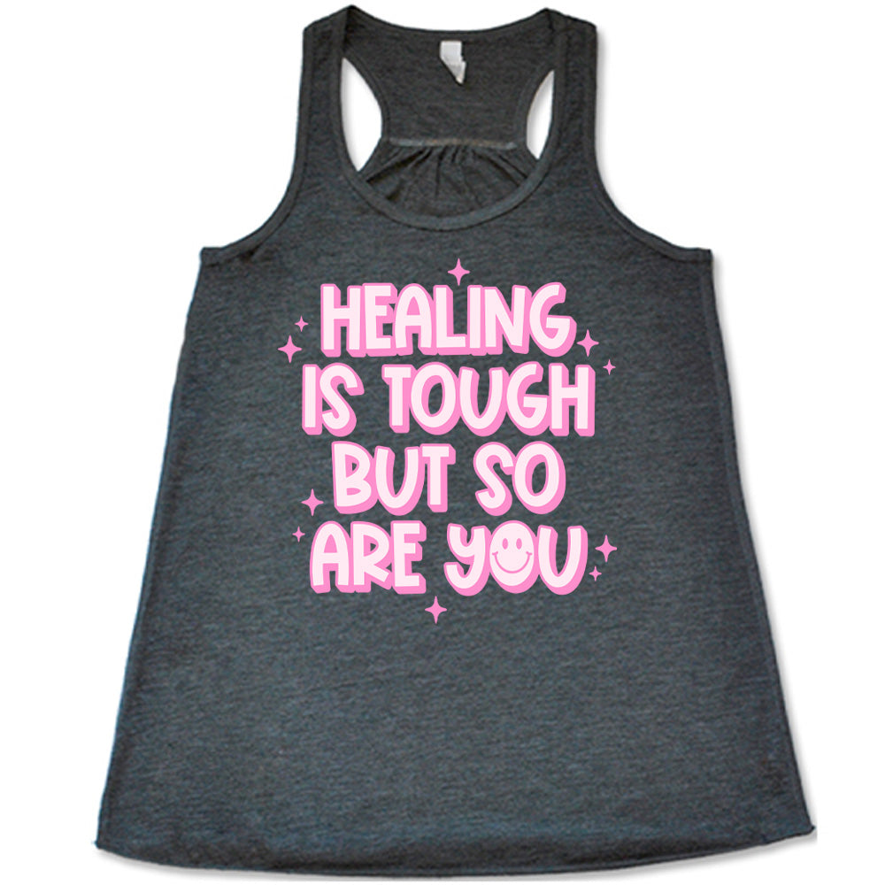 grey racerback flowy tank with the words "healing is tough but so are you" on it, with a smiley face in the word "you", and little star graphics scattered around