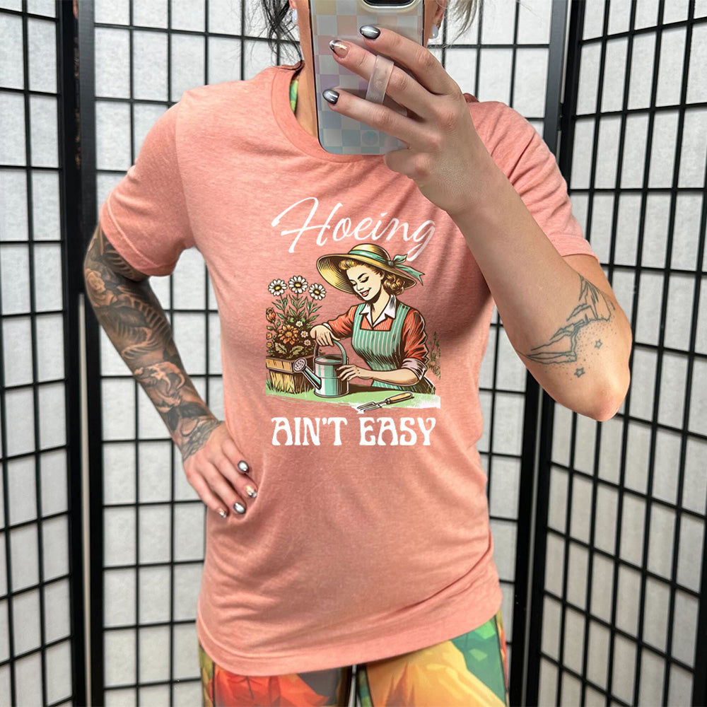 Model wearing sunset colored shirt with a woman gardening graphic on it that says "Hoeing Ain't Easy"