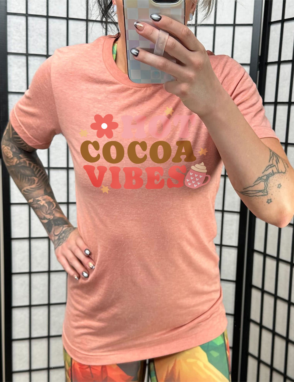 sunset unisex tee with the colorful words saying "HOT COCOA VIBES" with small graphics of a flower and mug, with many hearts on it, topped with whipped cream, and a few stars