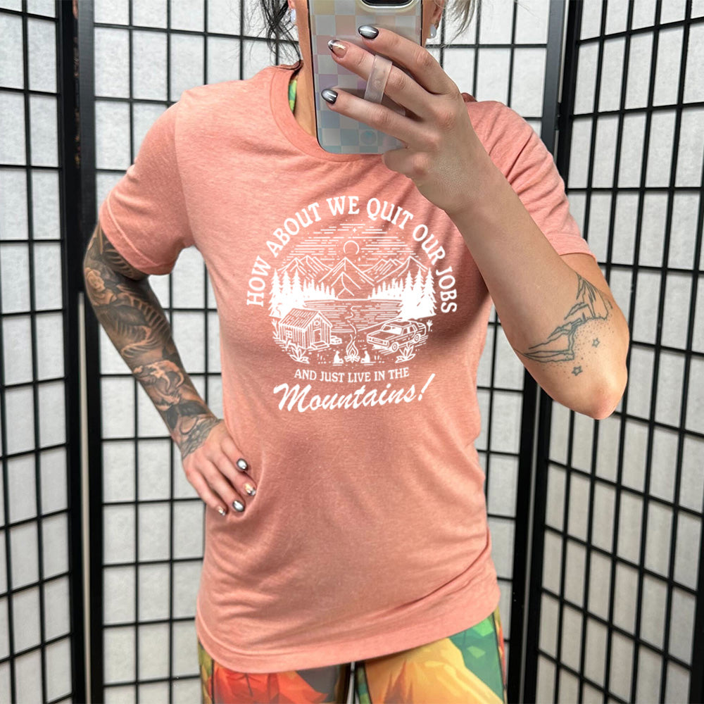 sunset unisex tee with the words "How about we quit our jobs and just live in the mountains!" on it, with the graphic of a small house, a couple people sitting around a campfire, and an old car in the woods and mountains, with a sky and a couple stars and the moon
