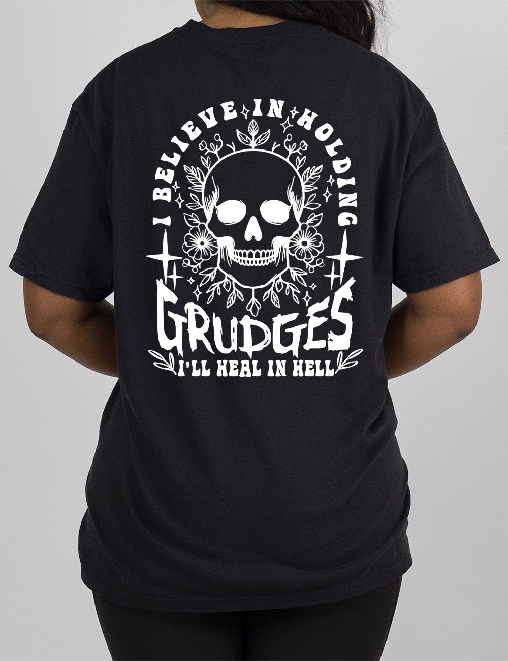 black modern tee with print placement on the back, with the graphic of a skull among flowers, and  words arching over it and words underneath, saying "I believe in holding GRUDGES I'll heal in hell"