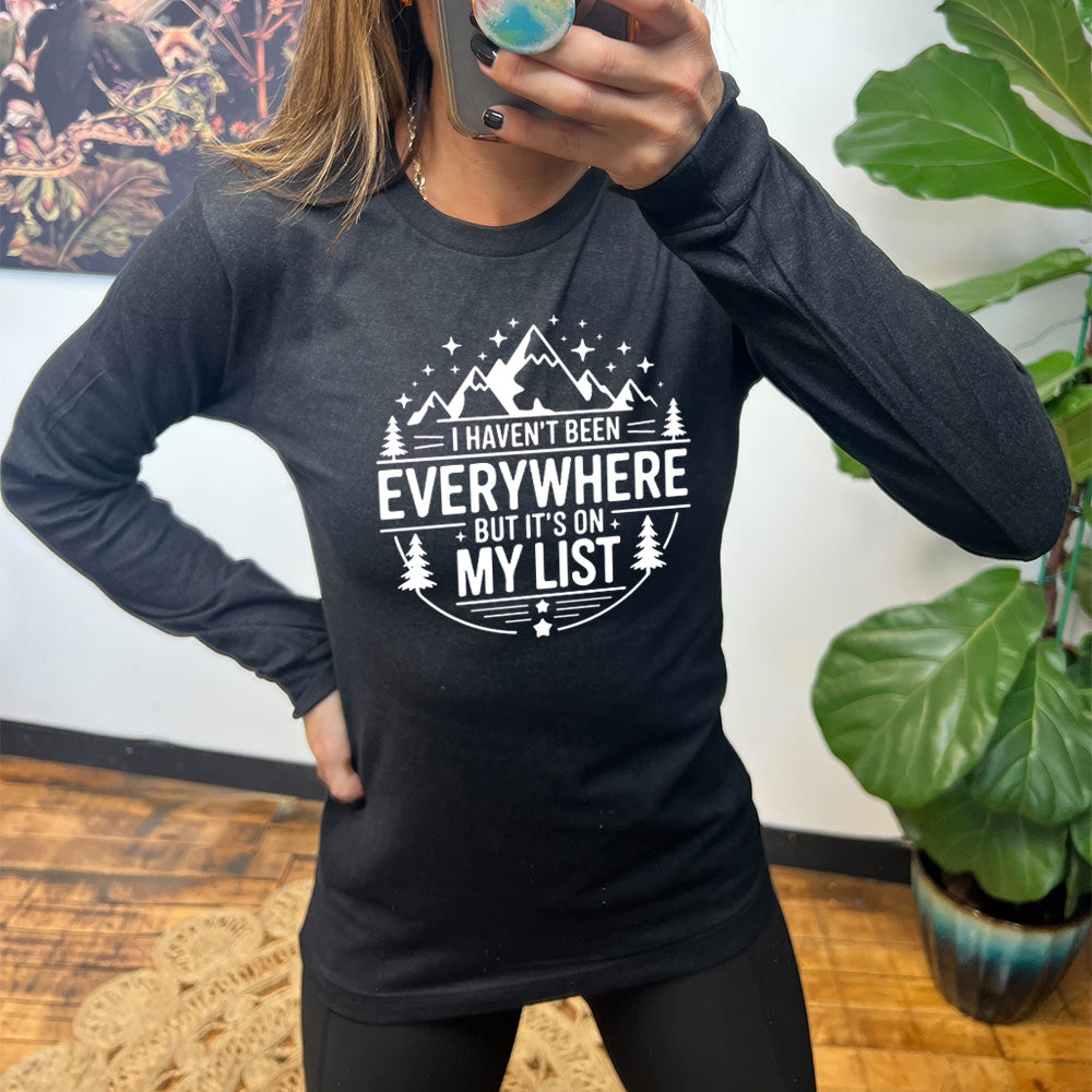 heather black long sleeve with the words "I haven't been everywhere but it's on my list" as part of a circular design involving the graphic of small mountains on top and a few trees on the sides and stars scattered around