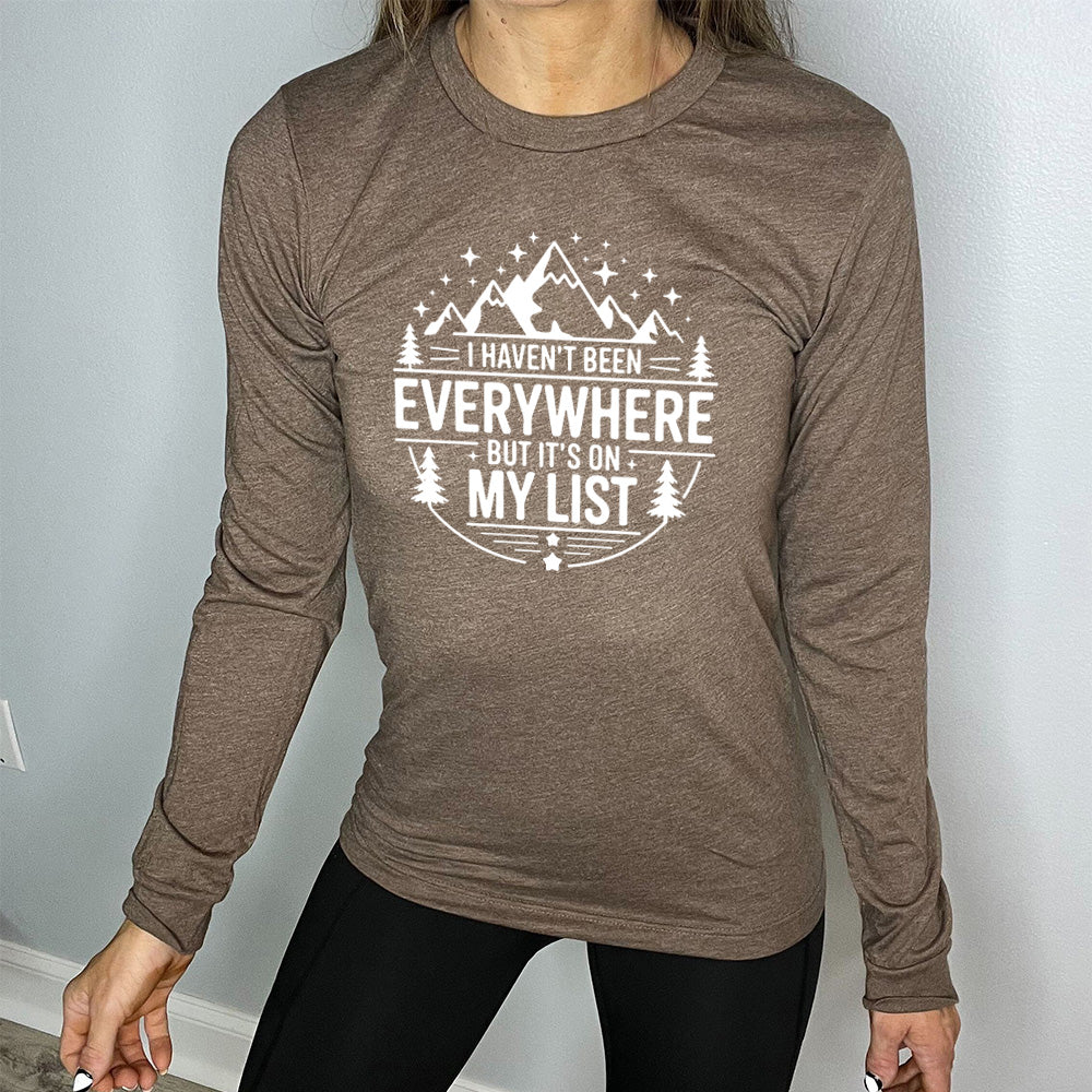 heather brown long sleeve with the words "I haven't been everywhere but it's on my list" as part of a circular design involving the graphic of small mountains on top and a few trees on the sides and stars scattered around