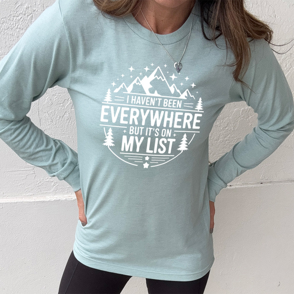 heather dusty blue long sleeve with the words "I haven't been everywhere but it's on my list" as part of a circular design involving the graphic of small mountains on top and a few trees on the sides and stars scattered around