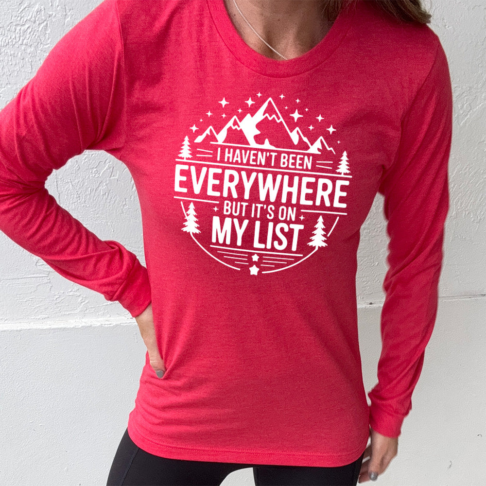 heather red long sleeve with the words "I haven't been everywhere but it's on my list" as part of a circular design involving the graphic of small mountains on top and a few trees on the sides and stars scattered around